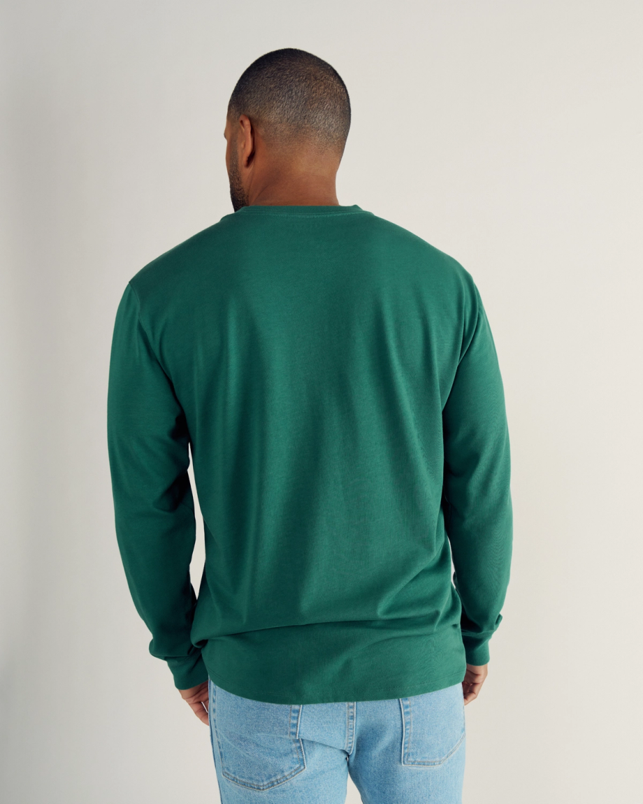 A man wearing a green long-sleeve shirt and light blue jeans stands facing away from the camera against a plain background.
