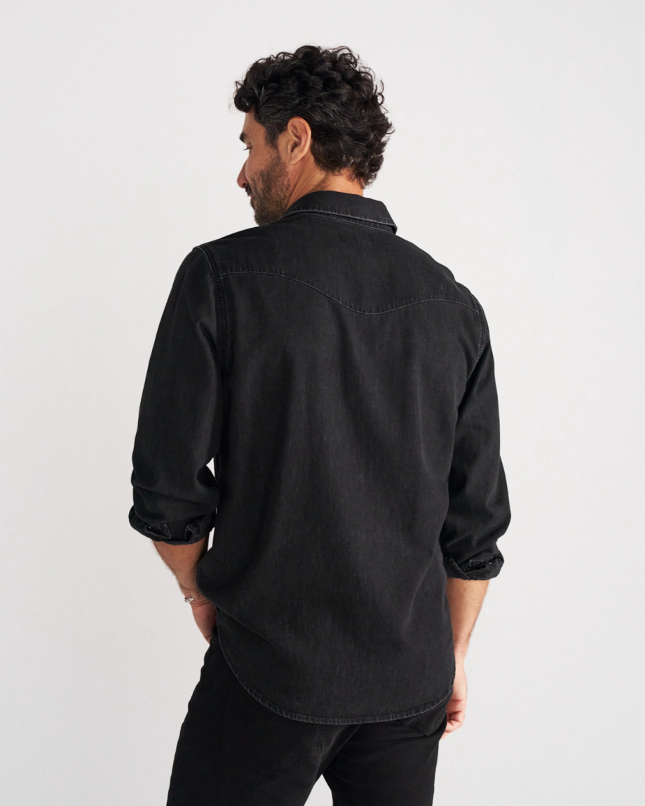 A man with dark curly hair is viewed from behind, wearing a black long-sleeved button-down shirt and black pants, standing against a plain white background.