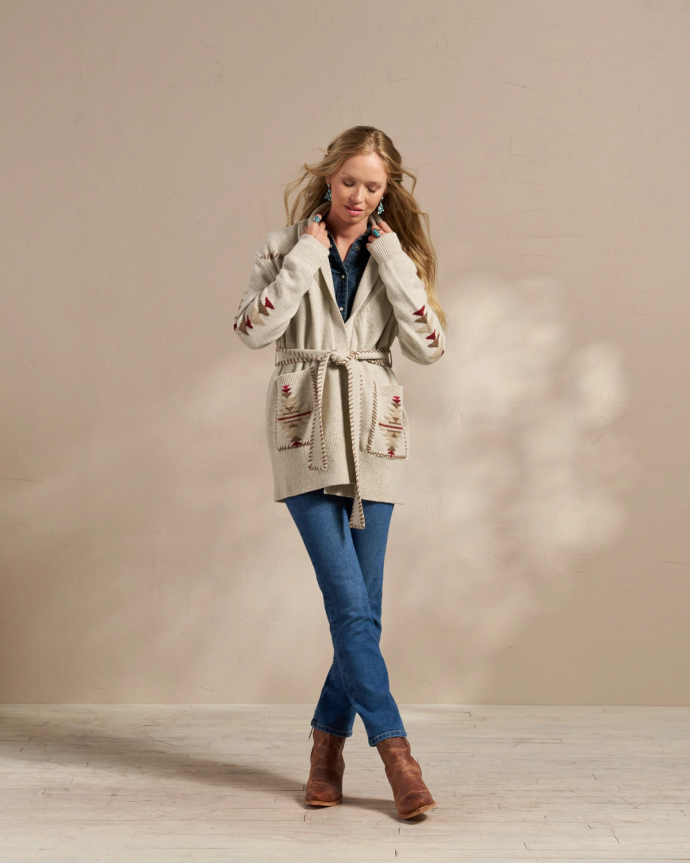 A woman stands against a plain beige background, wearing a belted beige cardigan, blue jeans, and brown boots, with her hands adjusting her collar.