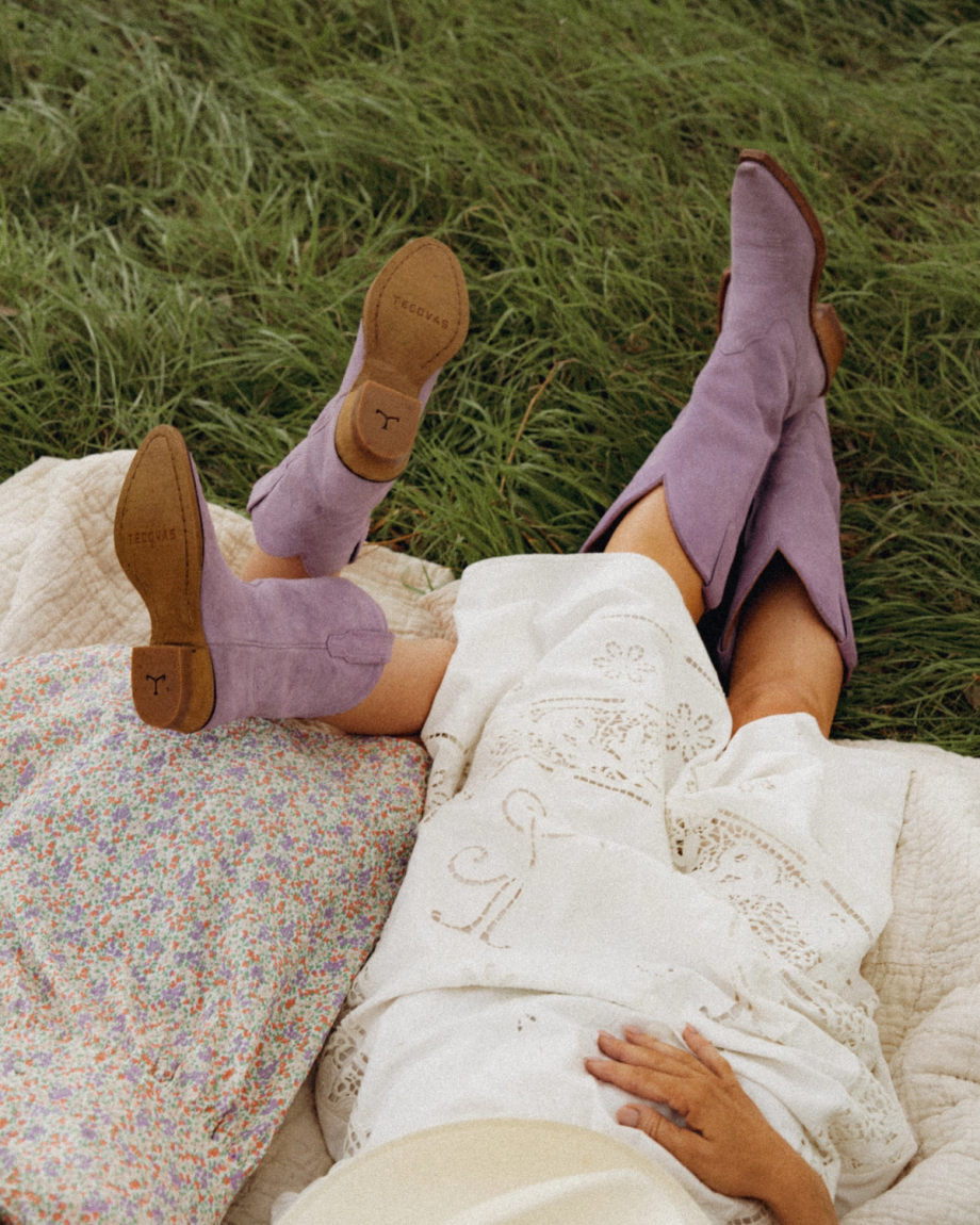 The Annie in Lavender Suede. Two people lie on a blanket in the grass, wearing light-colored skirts and stylish lavender suede cowboy boots, their legs extended and the boots beautifully visible.
