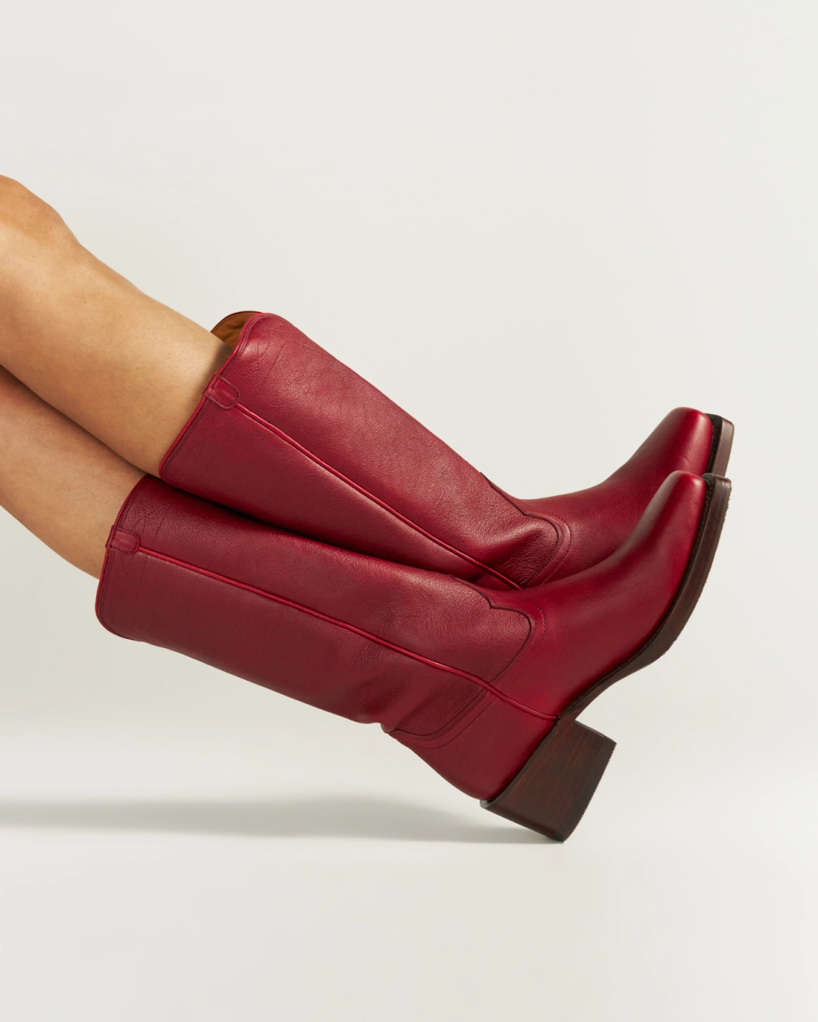The Charlie in Scarlet Cowhide. A person wearing tall, scarlet red leather cowboy boots with block heels—crafted in the signature Charlie Cowhide style—legs extended sideways against a plain white background.
