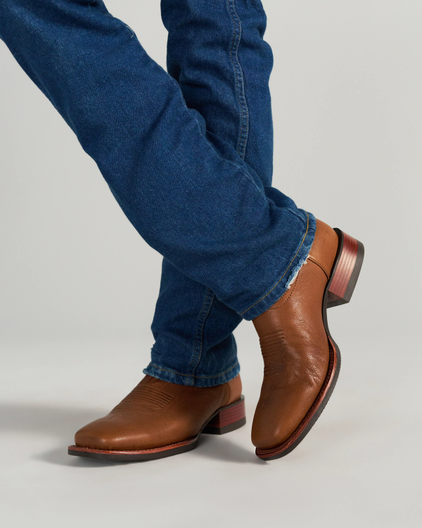The Dusty in Mesa Goat. Person wearing blue jeans and brown leather cowboy boots stands with legs crossed on a plain background, evoking the rugged spirit of The Dusty Goat.