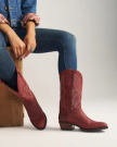 The Britt in Redwood Goat. A person wearing a denim jacket, jeans, and striking red cowboy boots sits on a wooden box from The Britt Goat, hands resting on their knee.