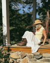 A woman in a yellow top, white skirt, and cowboy hat sits on a porch bordered by stone, with a metal chair and table in the background, surrounded by trees.