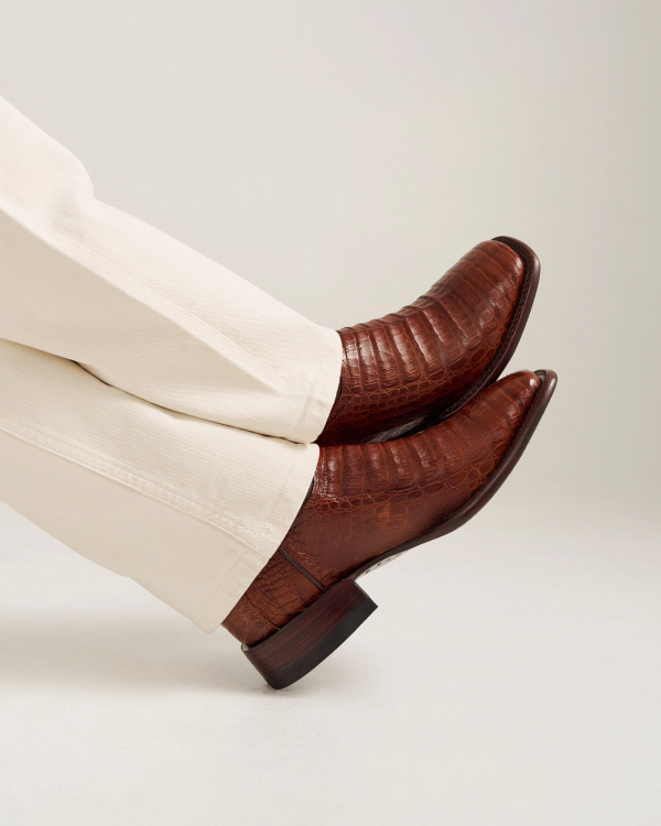 Person wearing off-white pants and Dark Pecan, crocodile-patterned leather boots, The William Caiman, with pointed toes, legs crossed at the ankles against a plain light background.