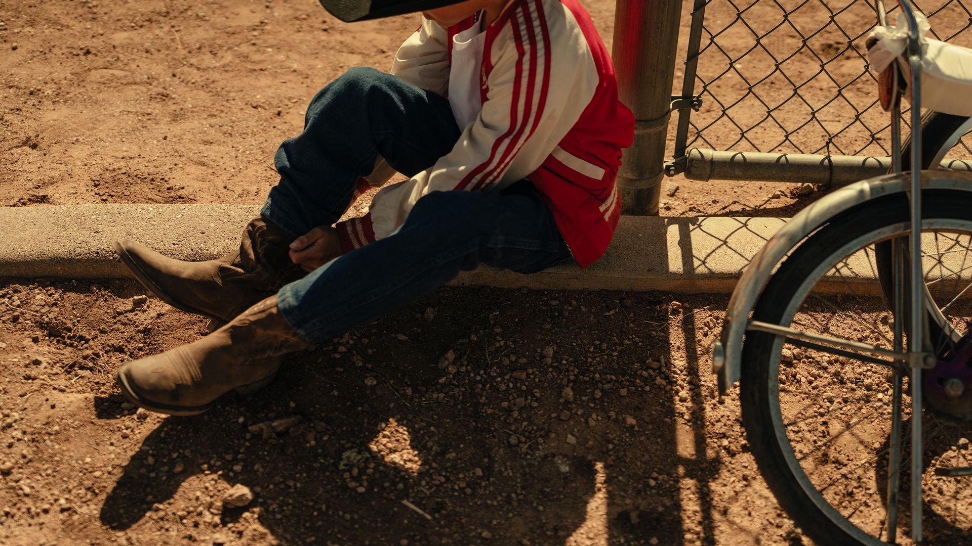 A child in a red and white jacket sits on the ground near a fence, putting on brown boots next to a bicycle.