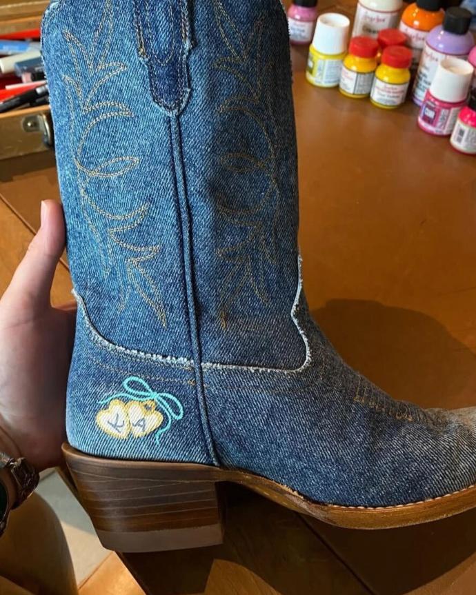 A hand holds a denim cowboy boot with a small, painted yellow chick and blue bow near the heel; art supplies are visible in the background.