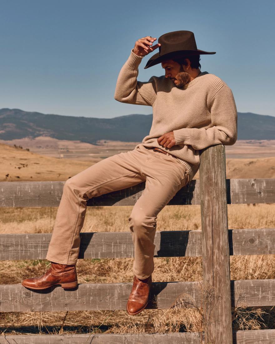 Man sitting on a wooden fence in a field, wearing a beige sweater, tan pants, brown boots, and a wide-brimmed hat against a clear blue sky.