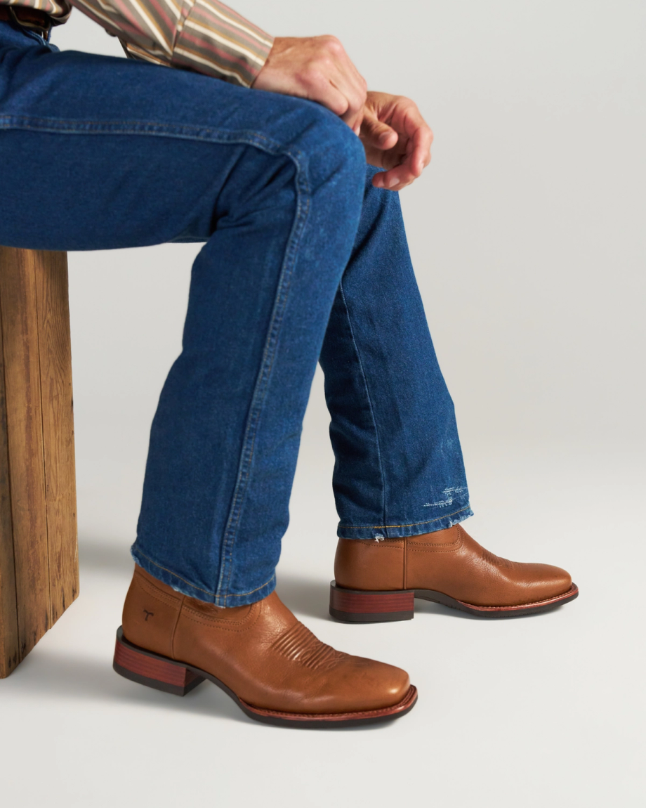 The Dusty in Mesa Goat. Person sitting on a wooden bench wearing blue jeans, a striped shirt, and brown leather ankle boots from The Dusty Goat, set against a plain background.