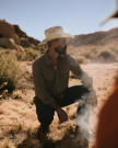 A man wearing a cowboy hat and button-up shirt kneels on dry grass in a desert landscape, with rocks and hills in the background.