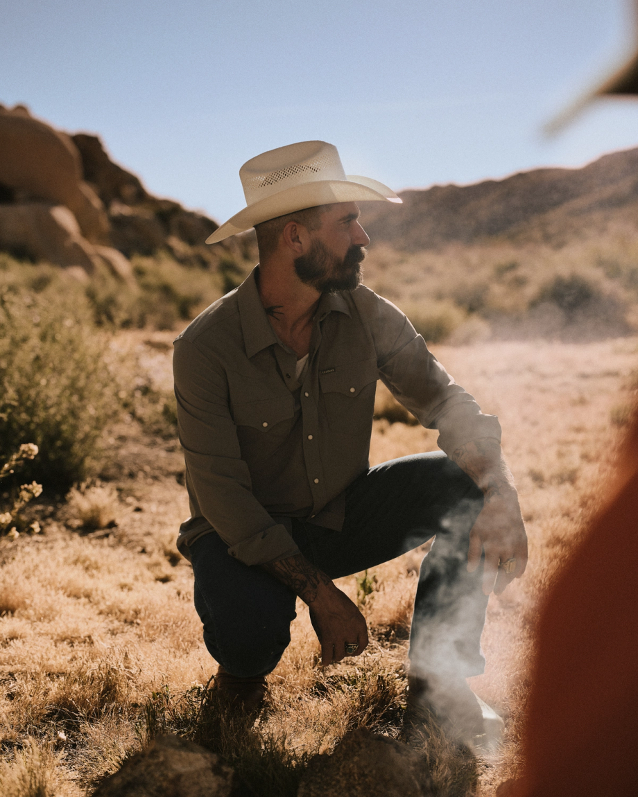 A man wearing a cowboy hat and button-up shirt kneels on dry grass in a desert landscape, with rocks and hills in the background.