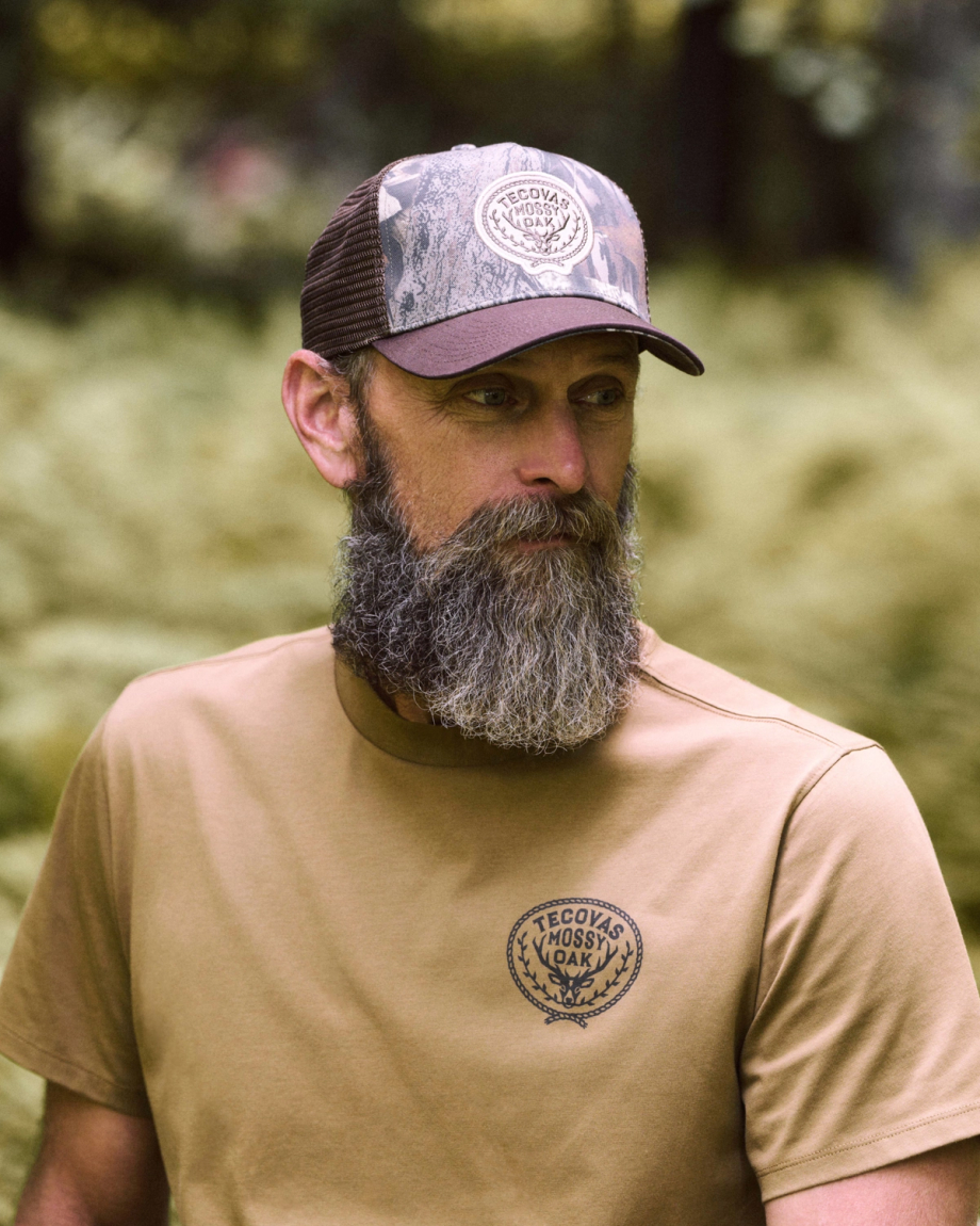 Man with a gray beard wearing a brown “Tecovas Mossy Oak” t-shirt and camouflage cap stands outdoors with greenery in the background.