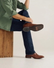 A person in a green jacket and blue jeans sits on a wooden block with legs crossed, wearing brown cowboy boots.