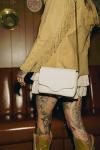 A person with tattooed legs stands in front of a leather couch, wearing a fringed tan jacket, white skirt, and holding a white handbag.
