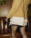 A person with tattooed legs stands in front of a leather couch, wearing a fringed tan jacket, white skirt, and holding a white handbag.