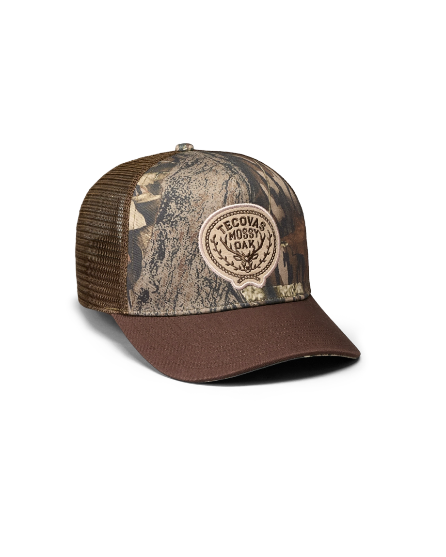 A brown and camouflage trucker hat with a mesh back and a round patch reading “Tecovas Mossy Oak” on the front.