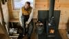 Man putting on boots inside a log cabin next to a wood burning stove