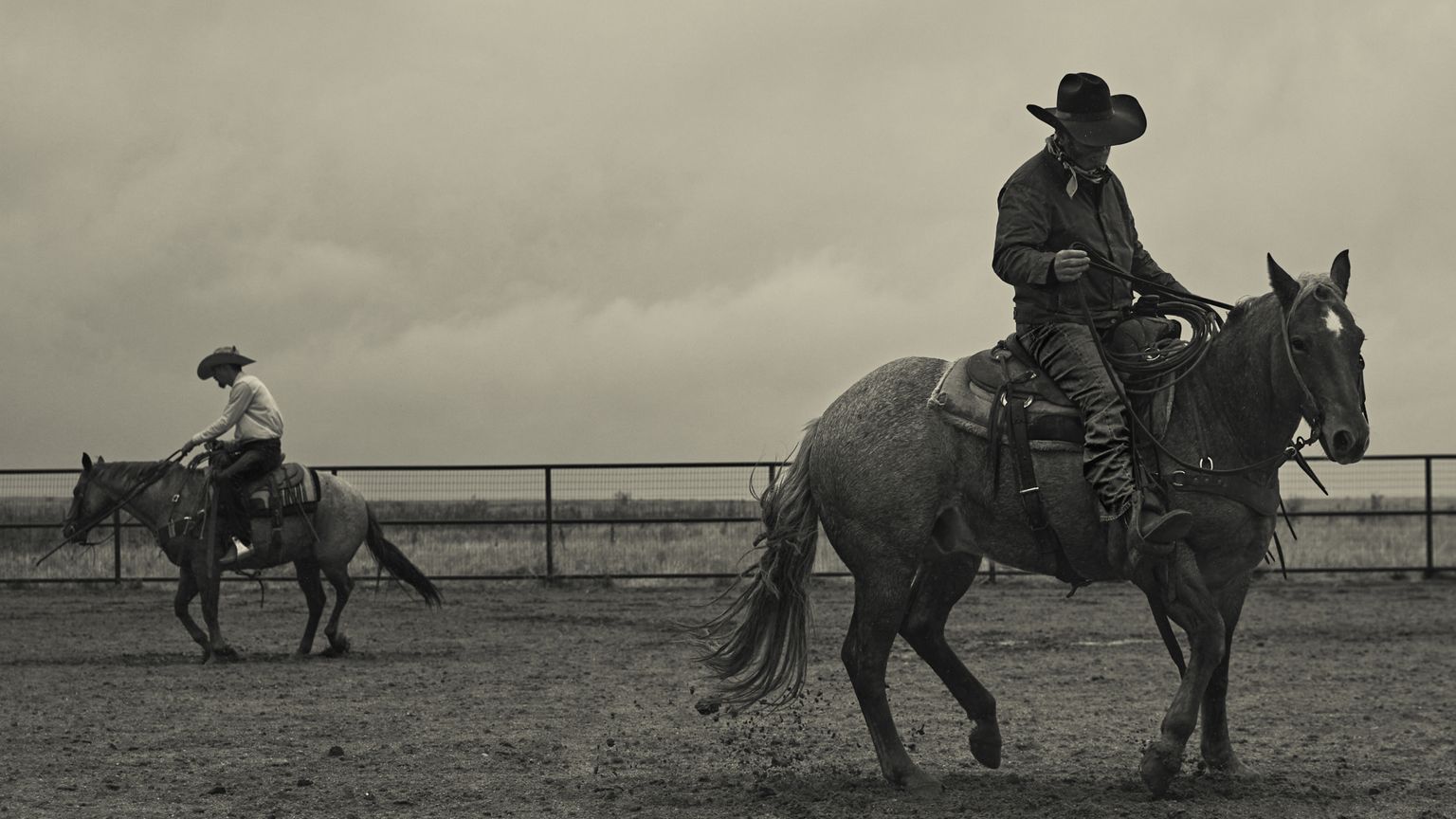 Two people on horseback in an enclosed arena, one in the foreground riding left, the other in the background moving right. The scene is set on a cloudy day.