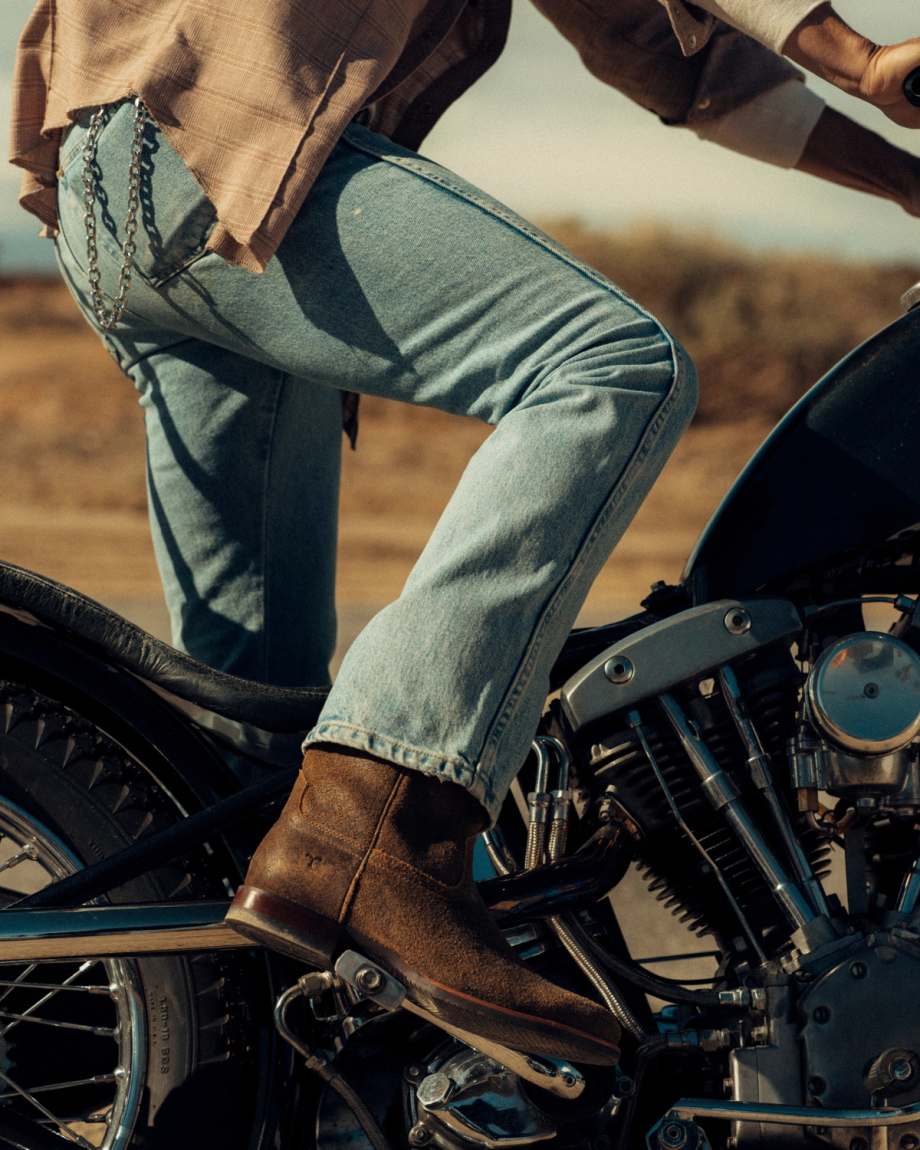 The Glen in Dustwood Roughout. Person wearing light blue jeans and brown boots sits on a vintage motorcycle, The Glen Roughout boots perfectly complementing the rugged desert landscape in the background.