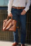 A person wearing a white shirt, jeans, and brown belt holds a brown leather briefcase. Only the lower half of the body is visible.