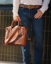 A person wearing a white shirt, jeans, and brown belt holds a brown leather briefcase. Only the lower half of the body is visible.
