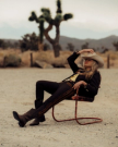 A person in a cowboy hat and western suit sits on a rusted chair in a desert landscape with Joshua trees behind, gazing thoughtfully as The Tess Goat cafe sign peeks from the sun-baked distance.