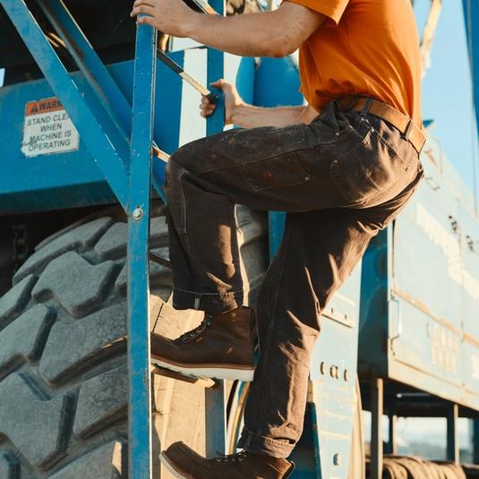 A person in an orange shirt and dark pants climbs a ladder attached to a large blue industrial machine with oversized tires.