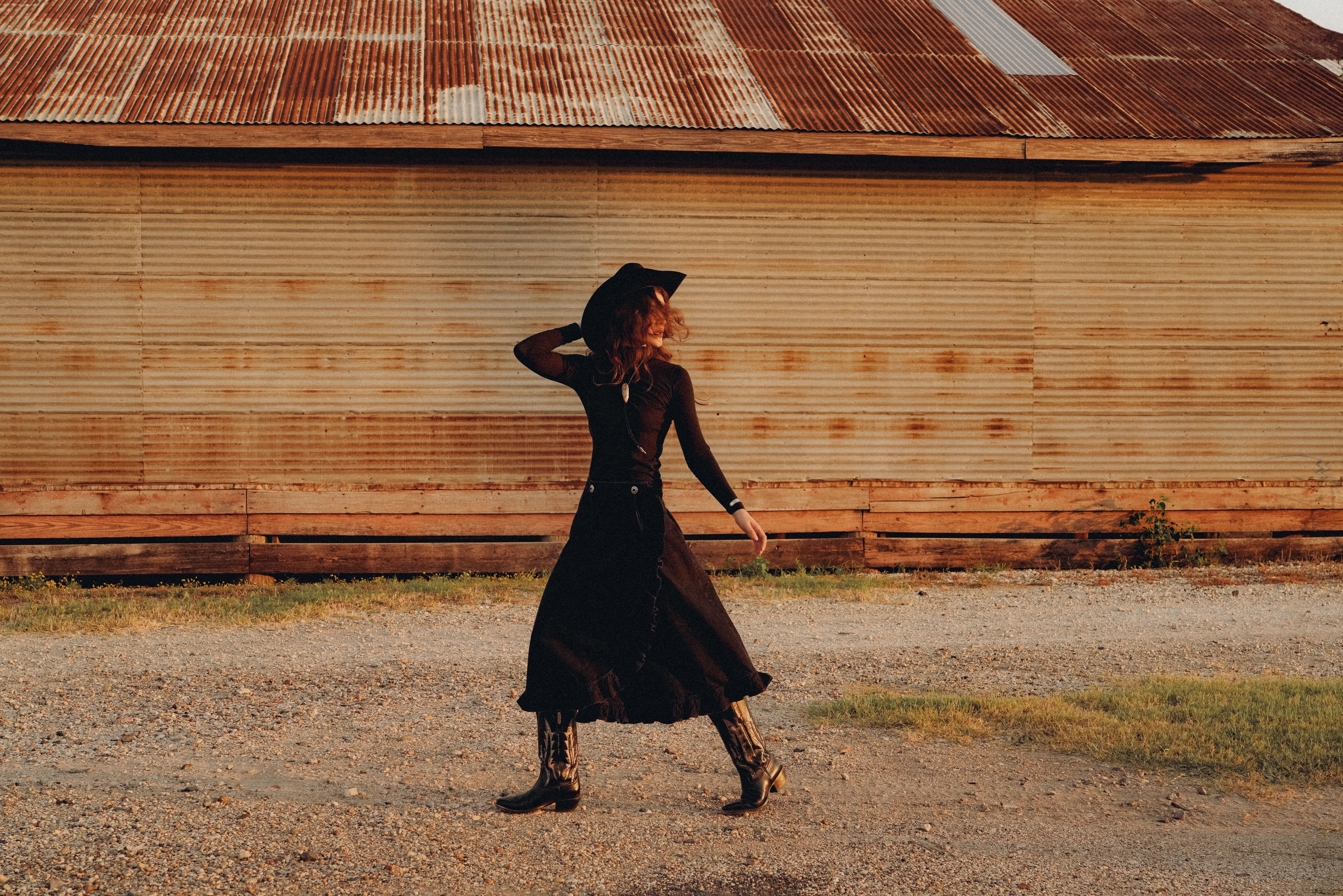 A person in a long black dress, hat, and boots walks across gravel in front of a corrugated metal building with a rusted roof.