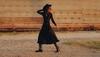 A person in a long black dress, hat, and boots walks across gravel in front of a corrugated metal building with a rusted roof.