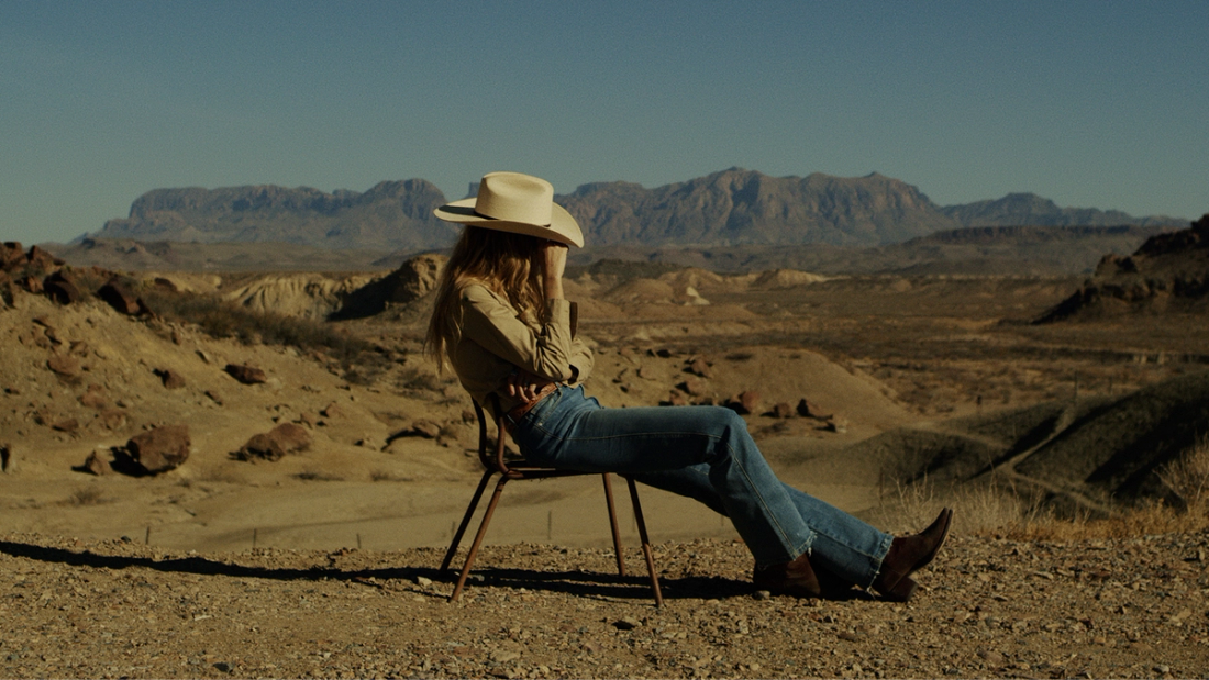 A person in a hat and jeans sits on a chair in a desert landscape, with mountains visible in the background under a clear sky.
