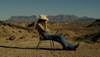 A person in a hat and jeans sits on a chair in a desert landscape, with mountains visible in the background under a clear sky.