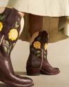 Dark brown leather cowboy boots with yellow rose and green leaf appliqués, worn with a layered skirt.