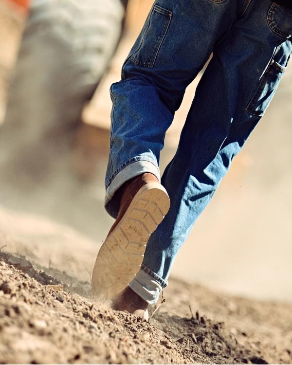 LongHaul Waterproof Roper Composite Toe Work Boot in Saddle Cowhide. A person in blue jeans and LongHaul Waterproof Roper Composite Toe Work Boot Cowhide walks on dry, dusty ground with a large vehicle tire visible in the blurred background.