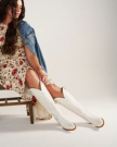 The Abby Wide Calf in Snow Cowhide. A person in a floral dress and denim jacket sits on a chair, wearing tall white cowboy boots inspired by The Abby Wide Calf Cowhide collection.