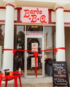 Front entrance of Barbs BQ restaurant with a red ribbon cutting ceremony set up, a sign on the door displaying the address as 102 E. Market St, and a chalkboard indicating where the line starts.