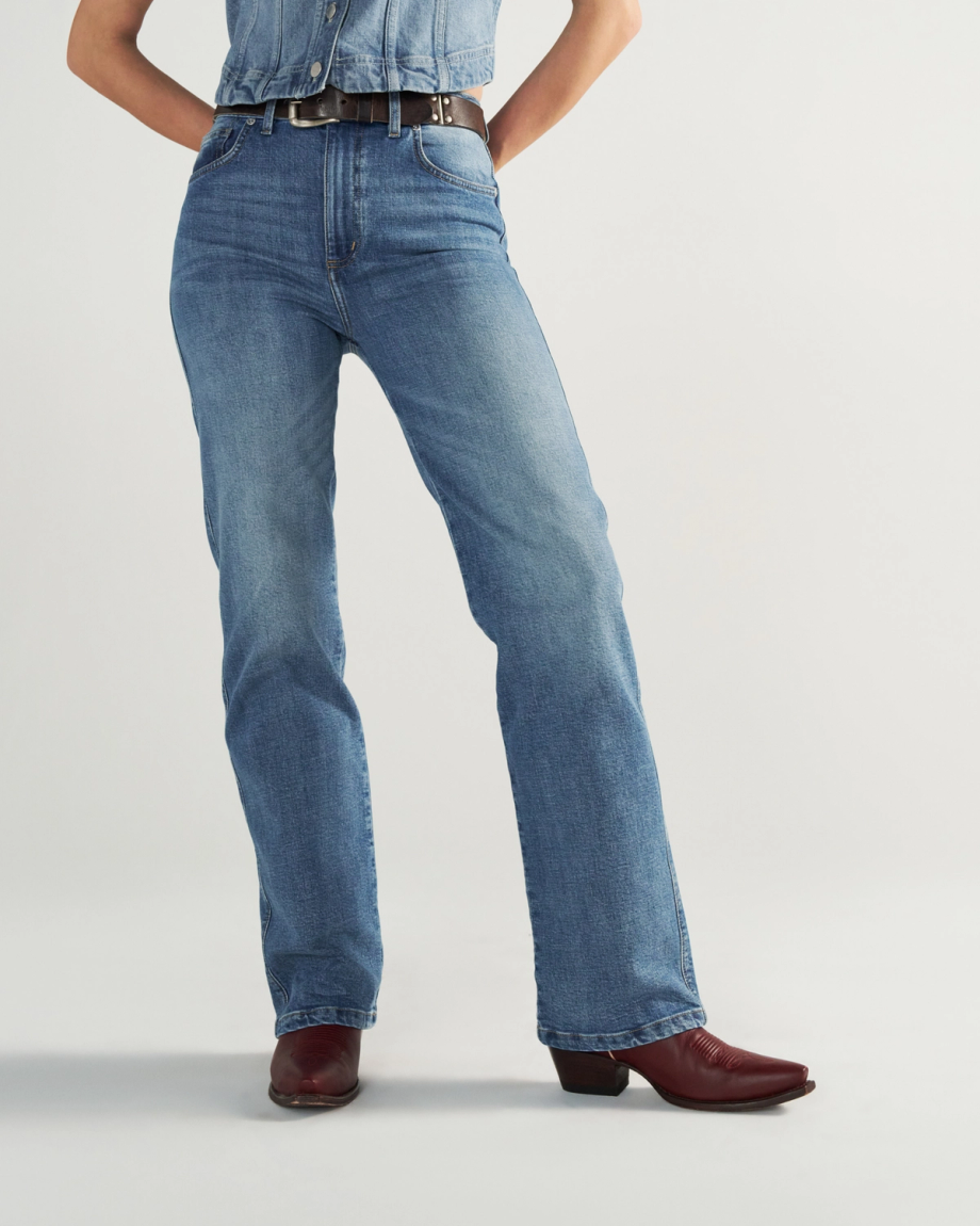 Person wearing blue jeans, a tucked-in denim shirt, a black belt, and burgundy cowboy boots, standing against a plain light background.