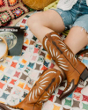 Person wearing brown cowboy boots and denim shorts, sitting on a patchwork quilt. There's a tambourine and a vinyl record nearby.