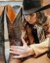 A woman in a cowboy hat and scarf carefully polishes a pair of tall leather boots on a table.