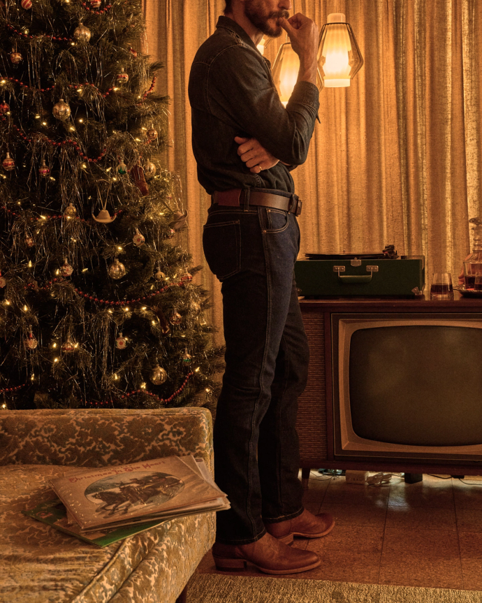 A man in jeans stands next to a vintage TV and record player in a warmly lit room with a decorated Christmas tree and wrapped gifts nearby.
