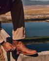 Person zipping brown leather boot, outdoors with blurred hilly landscape in the background.