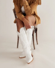 A woman stands against a plain background wearing a fringe jacket, white top, brown shorts, and white cowboy boots.