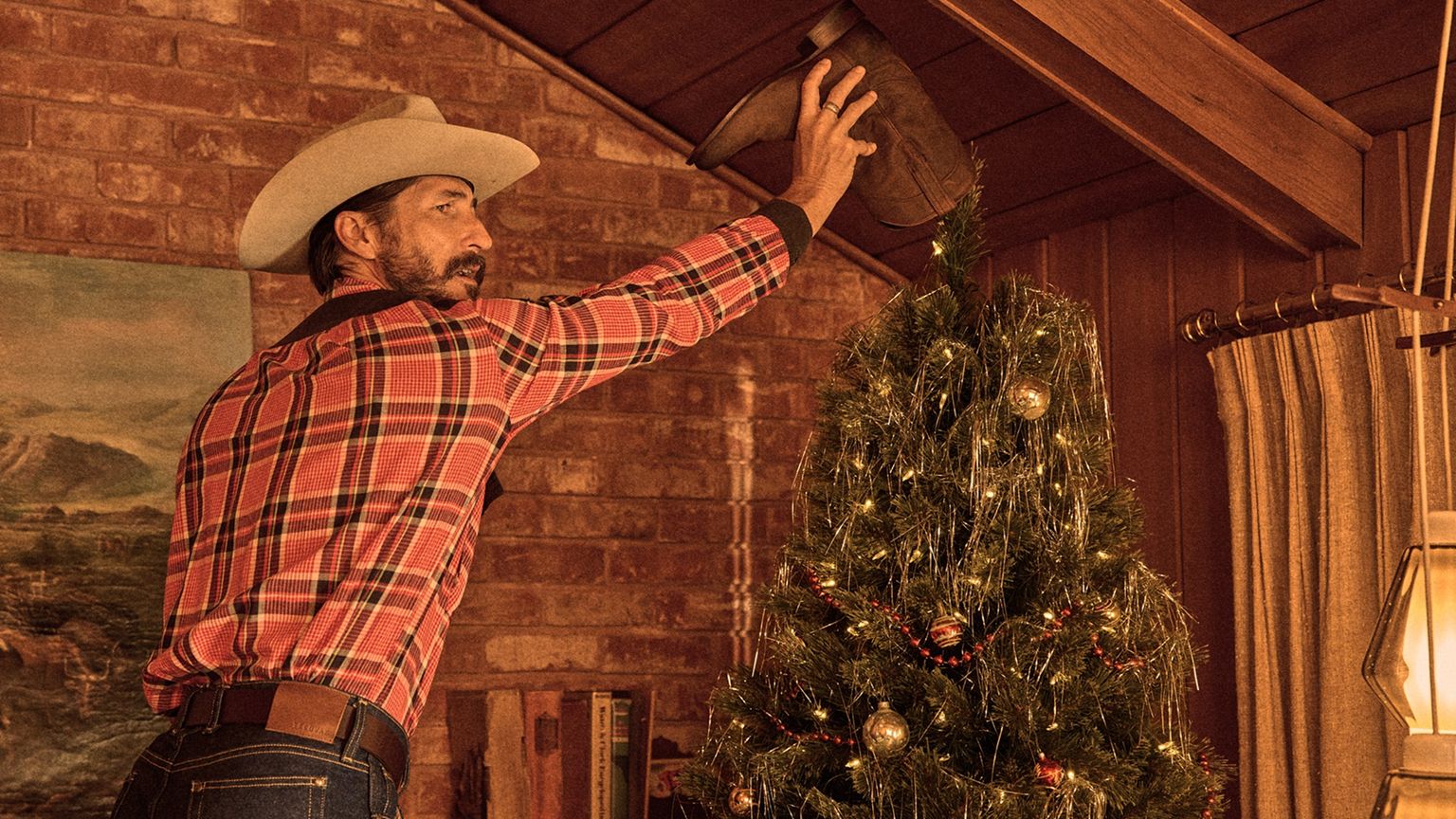 A man in a cowboy hat places a boot on top of a decorated Christmas tree in a wood-paneled room.