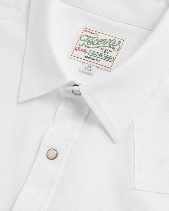 Close-up of a white collared shirt with a “Tecovas” brand label and pearl snap buttons, showing detail of stitching and fabric texture.