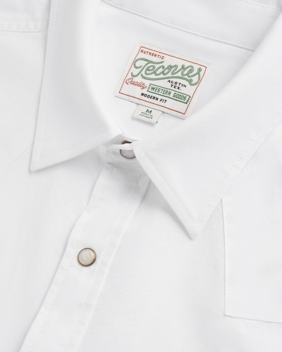 Close-up of a white collared shirt with a “Tecovas” brand label and pearl snap buttons, showing detail of stitching and fabric texture.