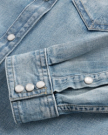 Close-up of a light blue denim shirt showing the cuff and part of the sleeve, with metal snap buttons and visible stitching details.
