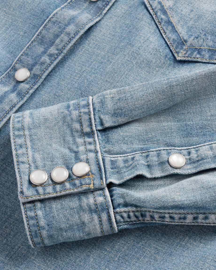 Close-up of a light blue denim shirt showing the cuff and part of the sleeve, with metal snap buttons and visible stitching details.