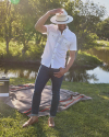 A man wearing a white shirt, jeans, and a hat stands on a grass field, covering part of his face with his hat. A blanket and a cooler are placed on the ground. Trees and a body of water are in the background.