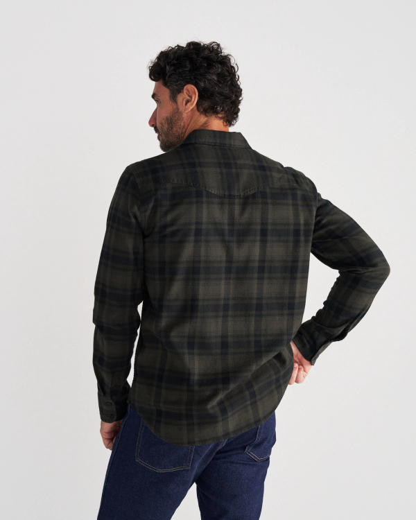 Man standing with his back to the camera, wearing a dark green and black plaid shirt and blue jeans against a plain white background.