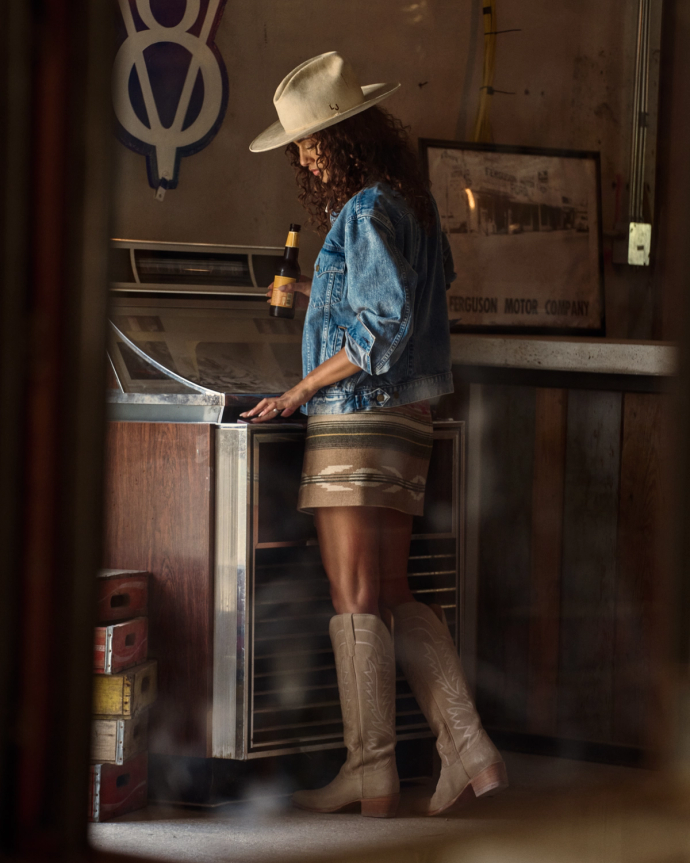 The Abby in Driftwood Suede. A woman in a hat, denim jacket, mini skirt, and The Abby Suede cowboy boots stands at a retro cooler, holding a bottle, in a rustic indoor setting.