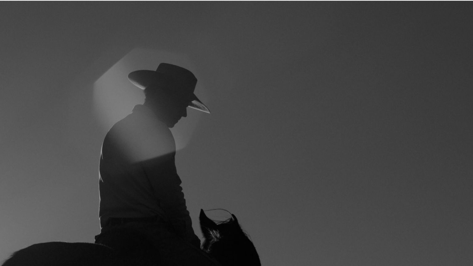 Silhouette of a person wearing a cowboy hat riding a horse, with the sun creating a bright halo effect in the background.